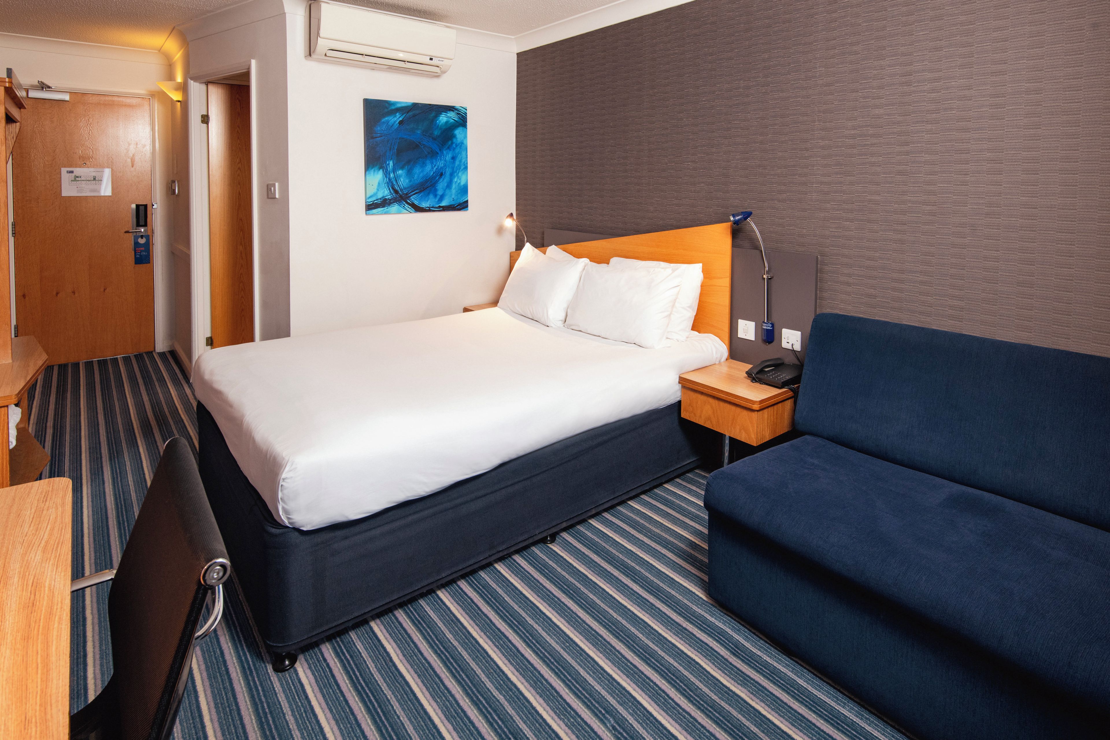 A hotel room with a bed, bedside table, lamp, and a blue sofa.