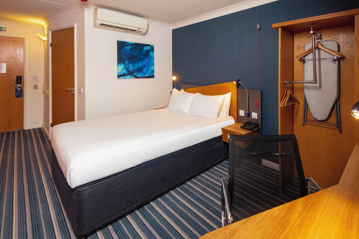 A hotel room with a bed, a desk, a chair, an ironing board, and a coat rack.