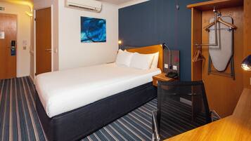 A hotel room with a bed, a desk, a chair, an ironing board, and a coat rack.
