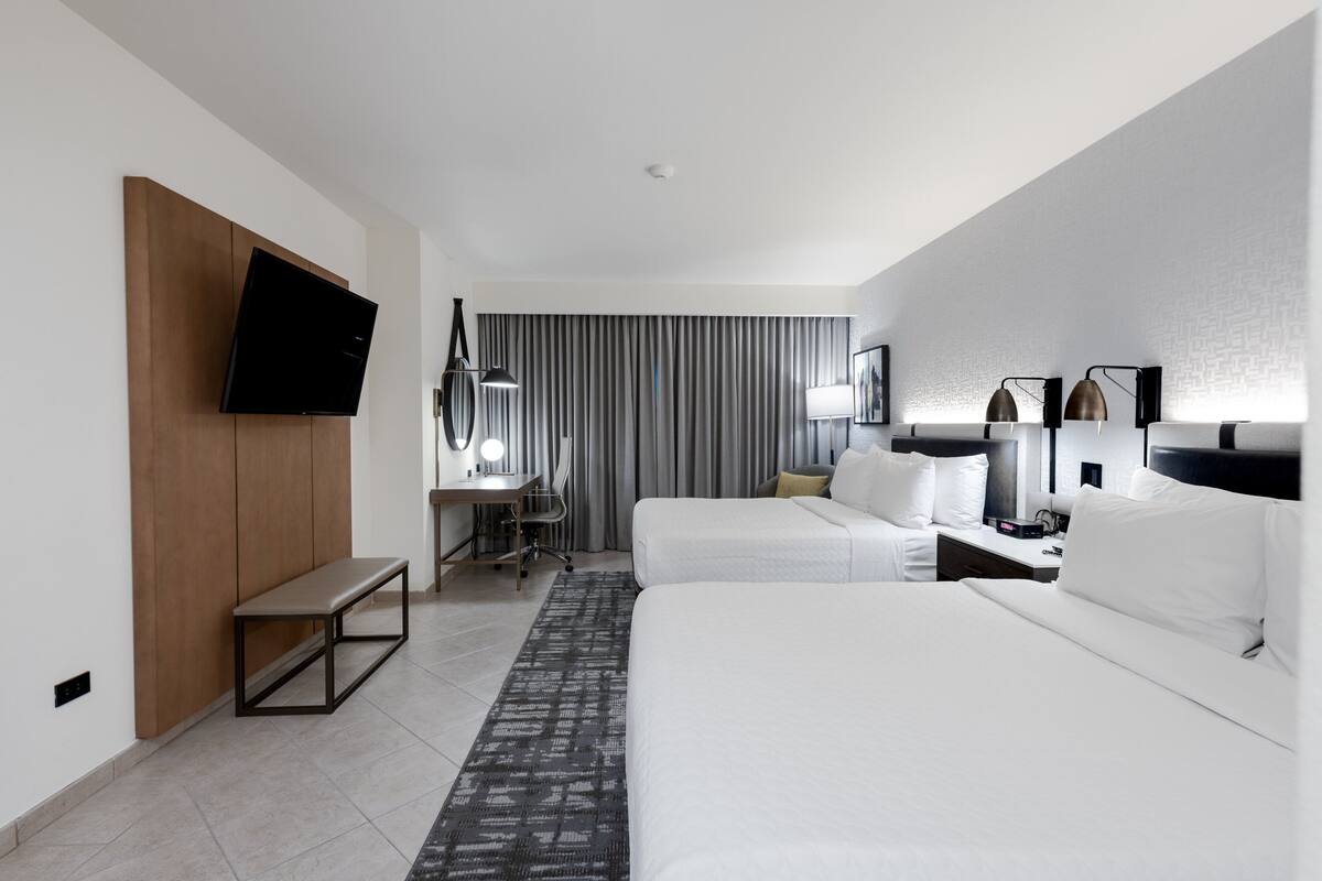 Una habitación de hotel moderna con televisor de pantalla plana montado en la pared, un escritorio con silla, cama con ropa de cama blanca y un ventanal con cortinas.