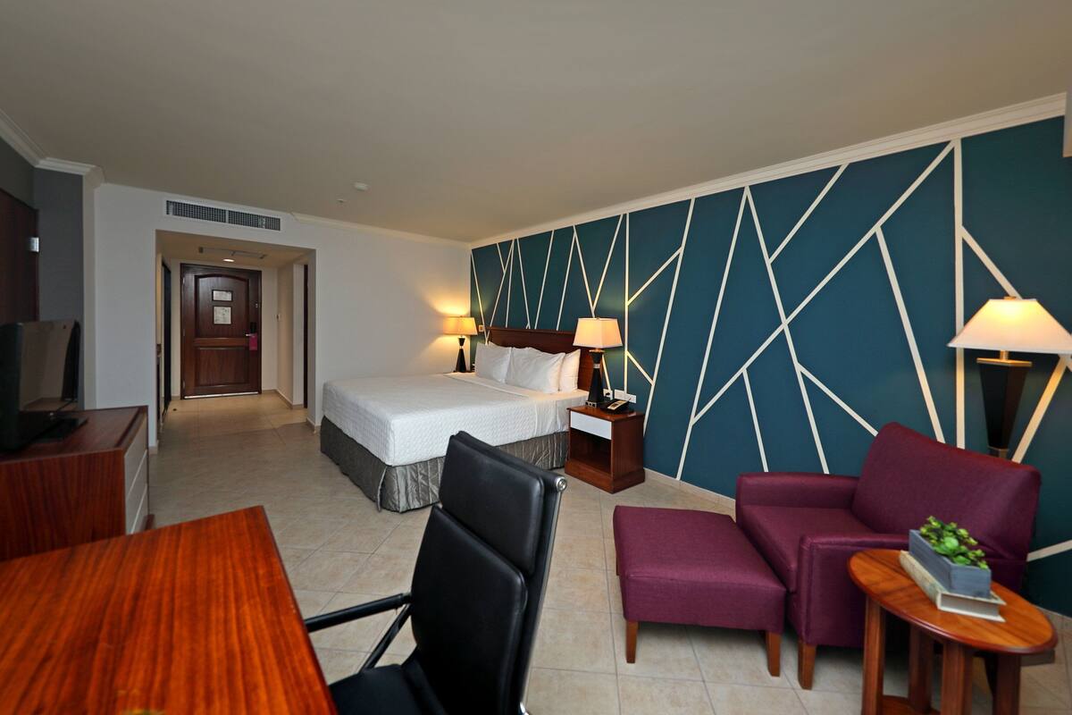 Habitación de hotel con cama, dos mesitas de noche, un escritorio, una silla y un sofá.