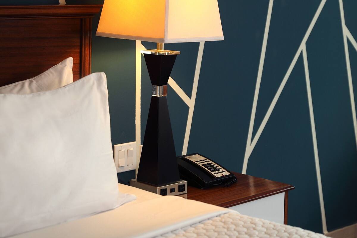 Una cama bien hecha con un almohadón blanco, una lámpara negra con pantalla blanca y un teléfono sobre una mesita de noche de madera. La pared presenta un diseño geométrico.