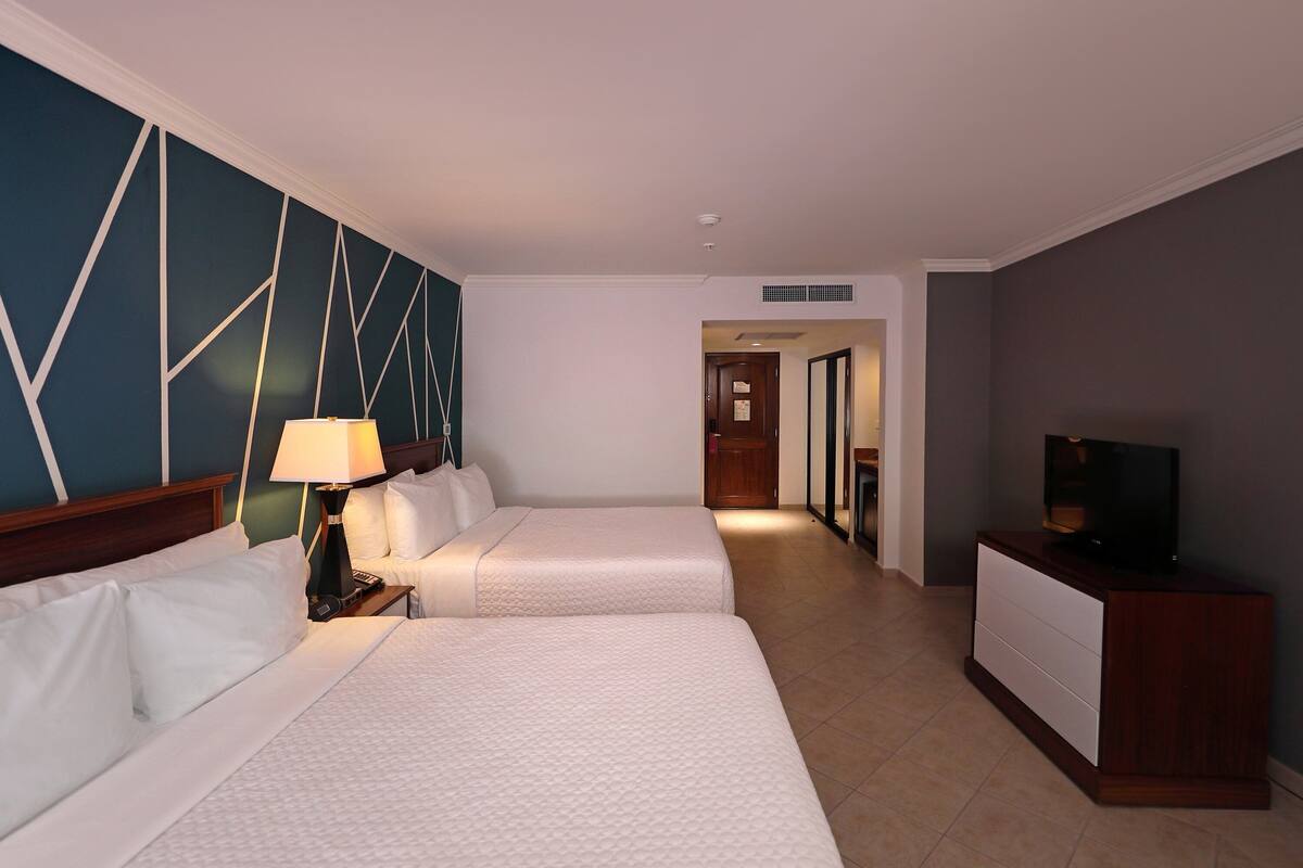 Habitación de hotel con dos camas, un televisor sobre un aparador y un pasillo que conduce a otra habitación.