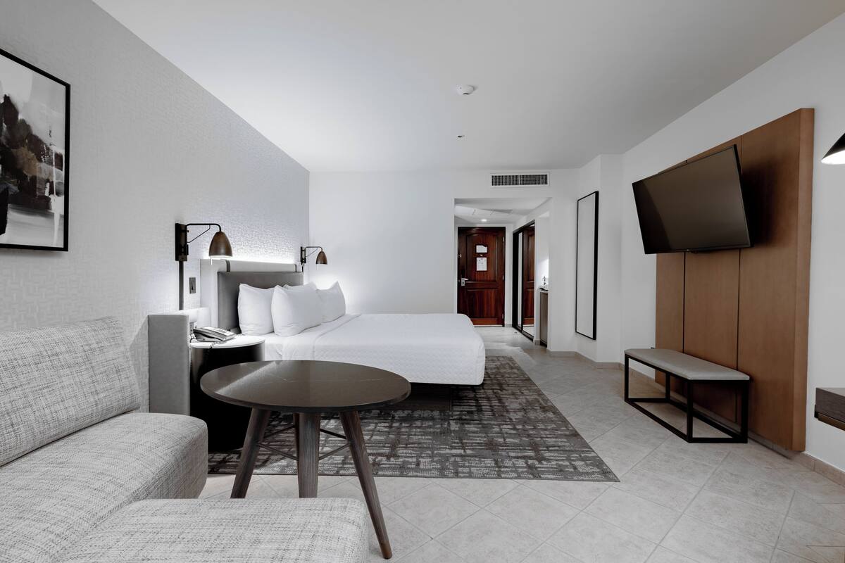 Una habitación de hotel moderna con una cama, un sofá, una mesita, un banco, un televisor y un cuadro en la pared.