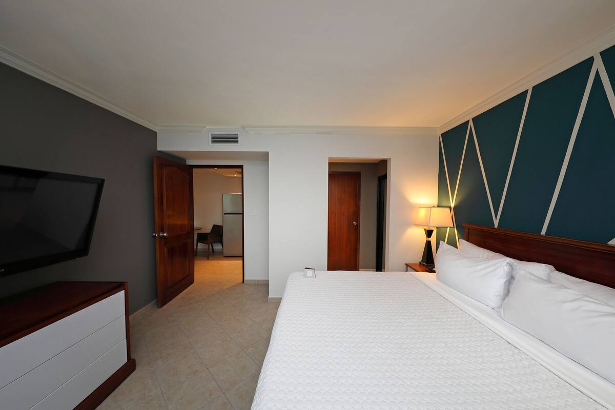 Habitación de hotel con una cama grande, un televisor en una cómoda, un escritorio con una lámpara y vistas al pasillo.