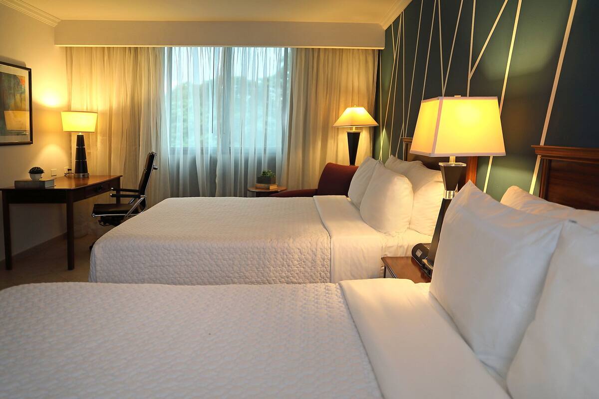 Habitación de hotel con dos camas, un escritorio, una silla, una lámpara y vistas al exterior a través de cortinas translúcidas.