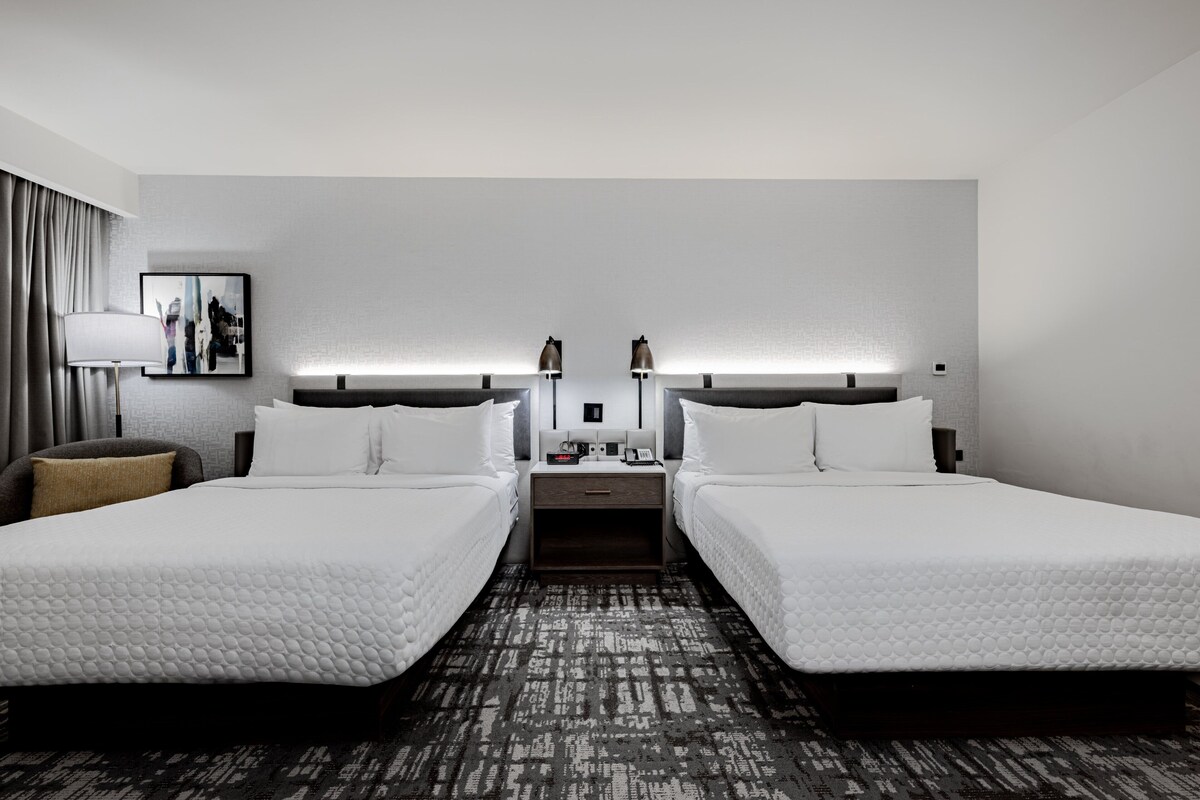 Habitación de hotel con dos camas individuales, una mesita de noche con lámparas, un pequeño tocador y un cuadro en la pared.