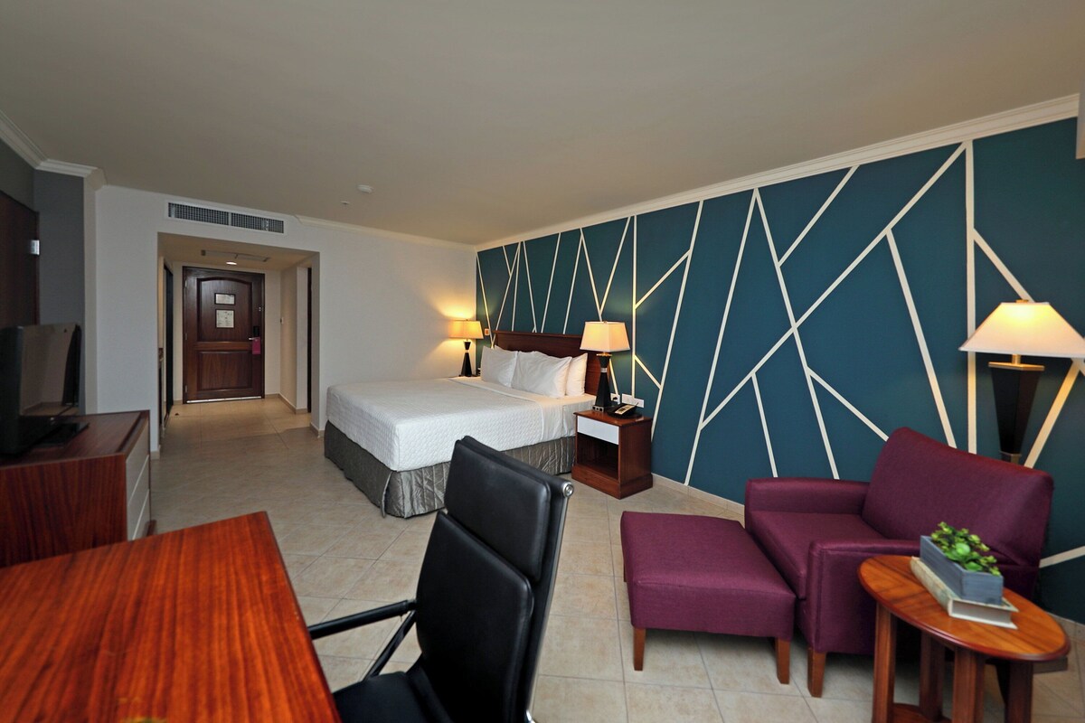 Habitación de hotel con una cama, dos mesitas de noche con lámparas, un escritorio con silla, un televisor y un área de descanso con sofá y sillones.