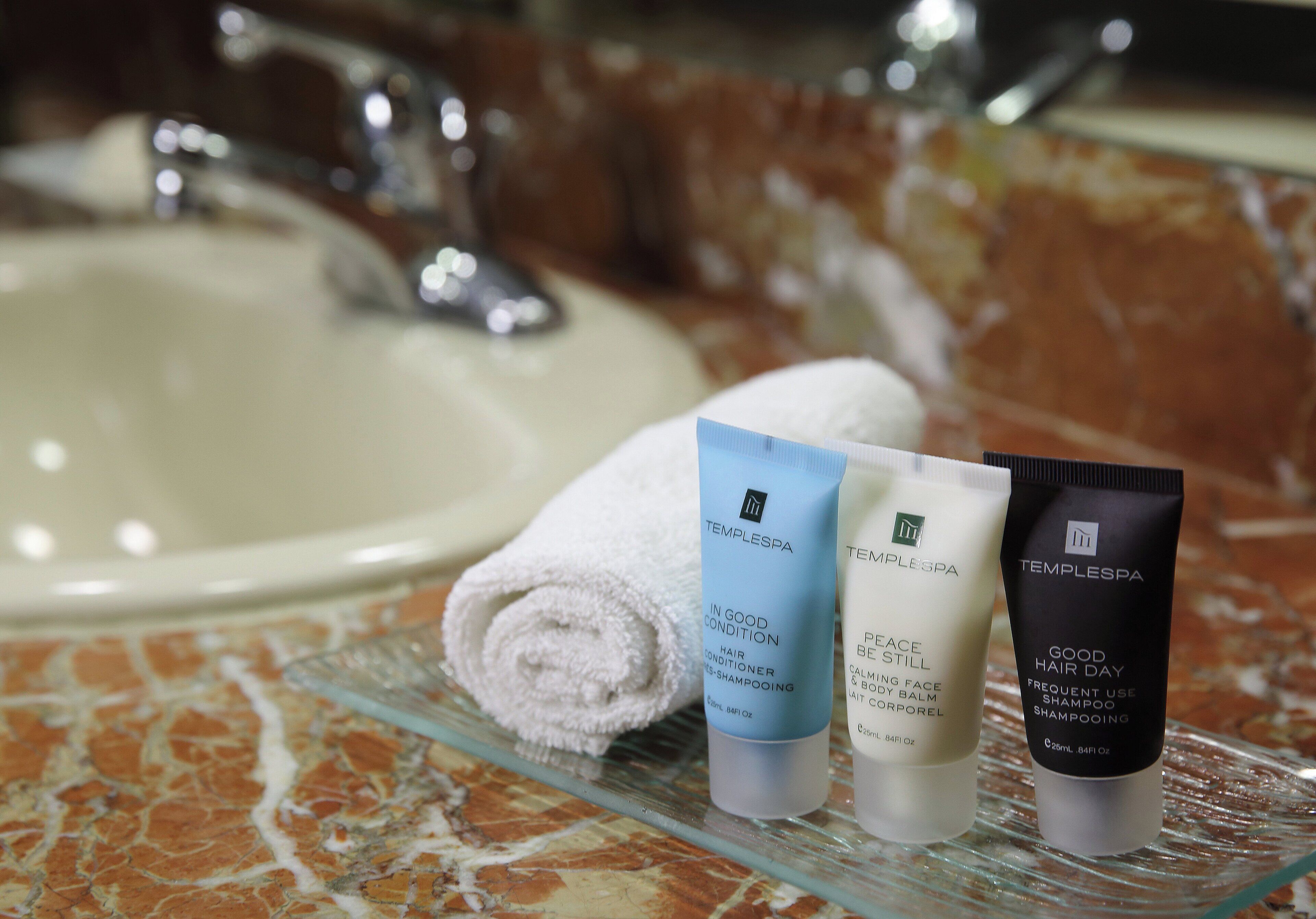 A bathroom with a marble sink, a glass shelf holding three toiletries, and a rolled white towel.