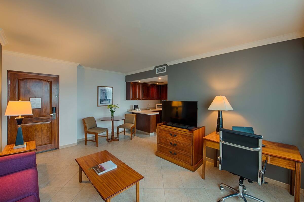 Habitación de hotel con escritorio de madera, televisión, zona de comedor con una mesa pequeña y sillas, y kitchenette.
