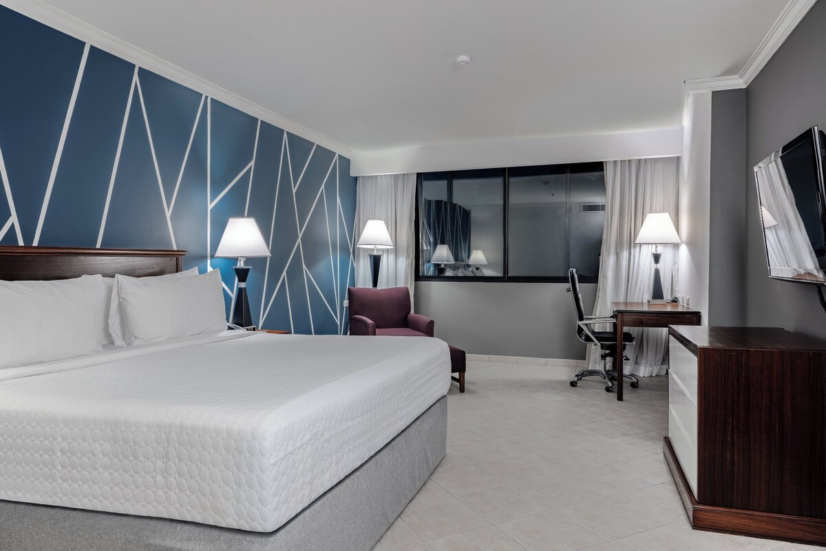 Habitación de hotel con una cama grande, dos mesitas de noche con lámparas, un escritorio con silla, un televisor y una ventana con cortinas.