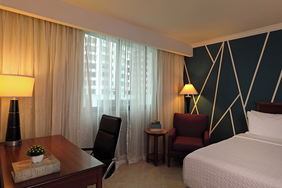 Habitación de hotel con cama, dos sillas, escritorio, lámpara y vista a un edificio a través de cortinas translúcidas.