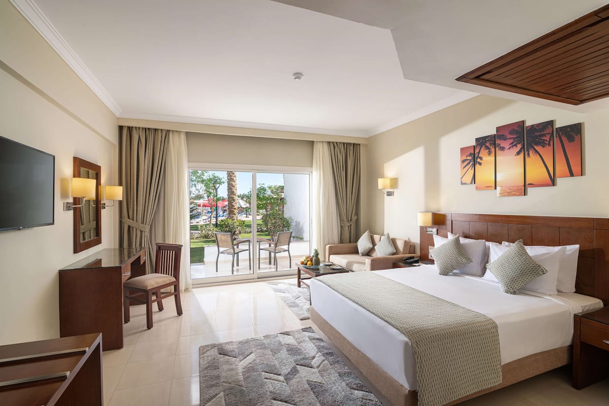 A hotel room with a large bed, a desk, a chair, a TV, and a balcony with a view of palm trees and a sunset.