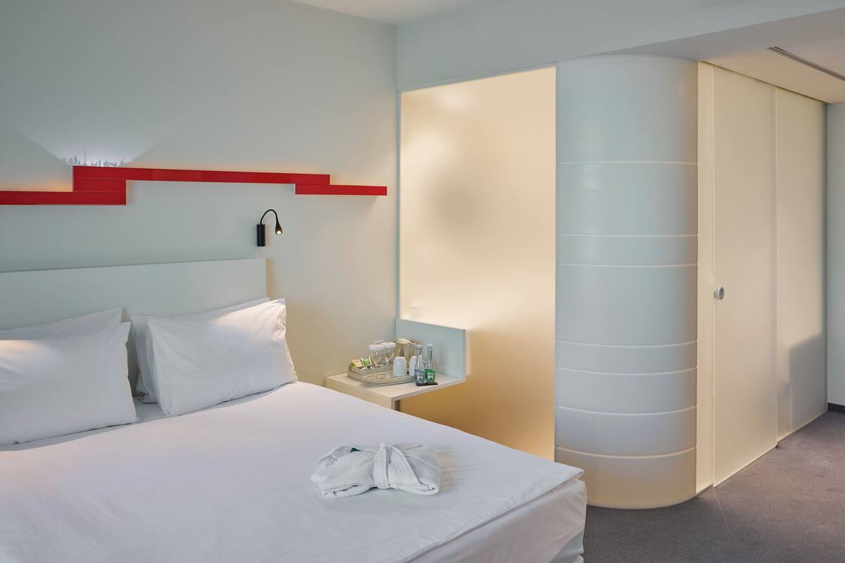 A hotel room with a bed, white pillows, a bedside table with a tray of toiletries, a robe, and a wardrobe with a light-colored door.