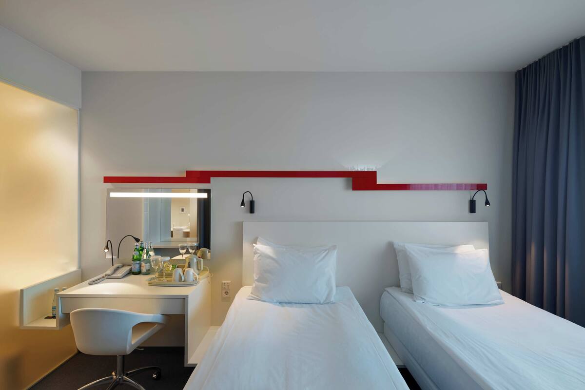 A hotel room with a desk, two beds, a chair, a mirror, and a red accent wall.