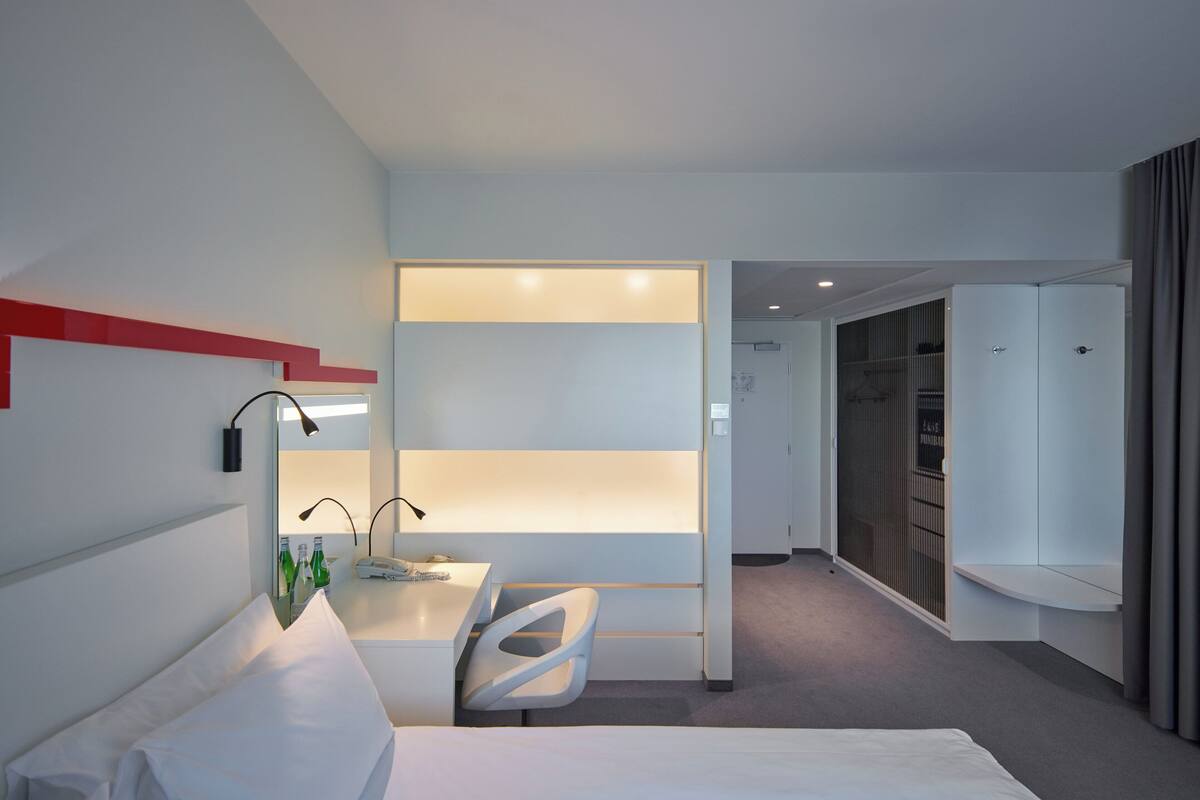 A modern hotel room with a bed, a desk with a chair, a minibar, and a walk-in closet.