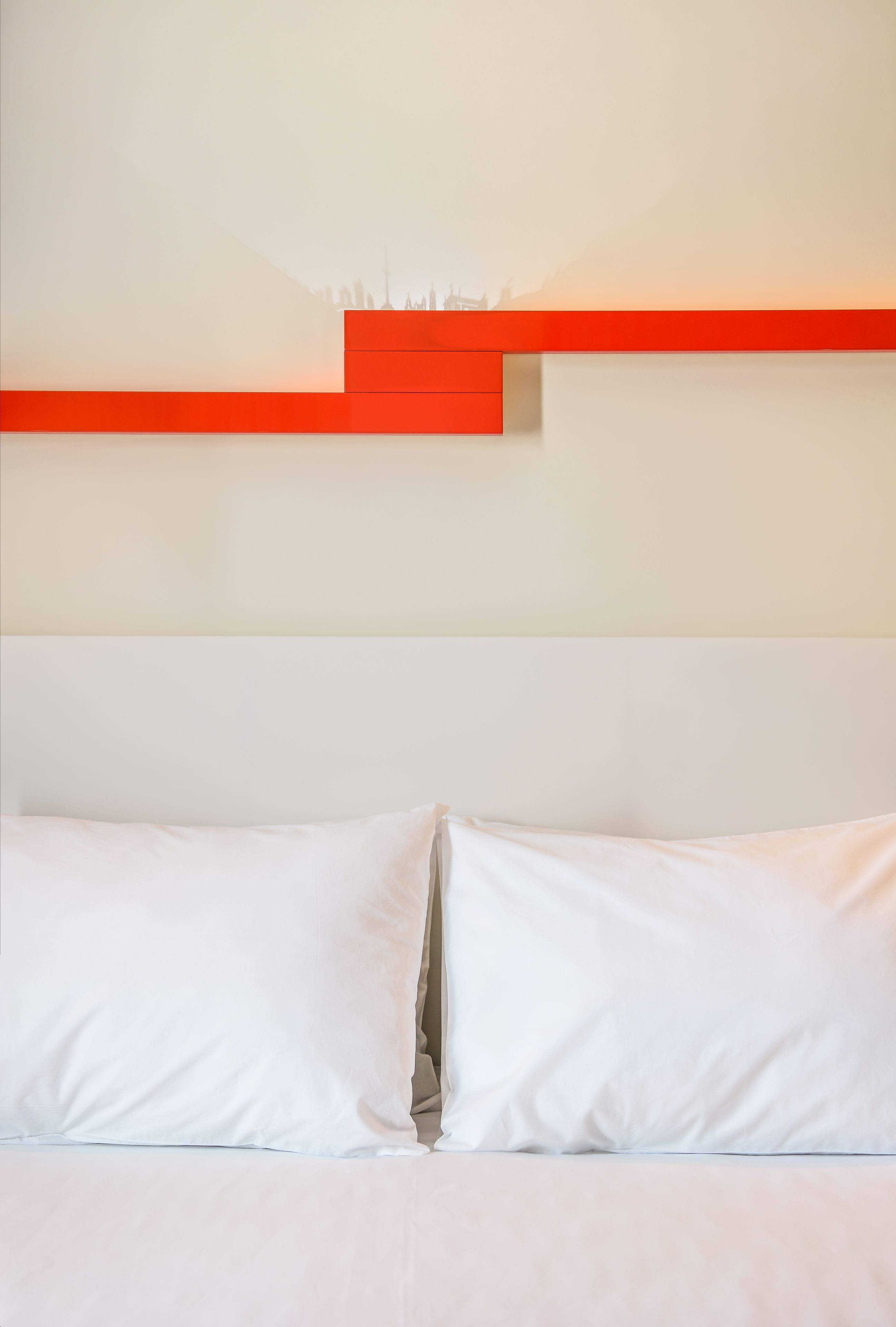 A bed with white bedding and pillows, a red accent wall, and a city skyline silhouette in the background.