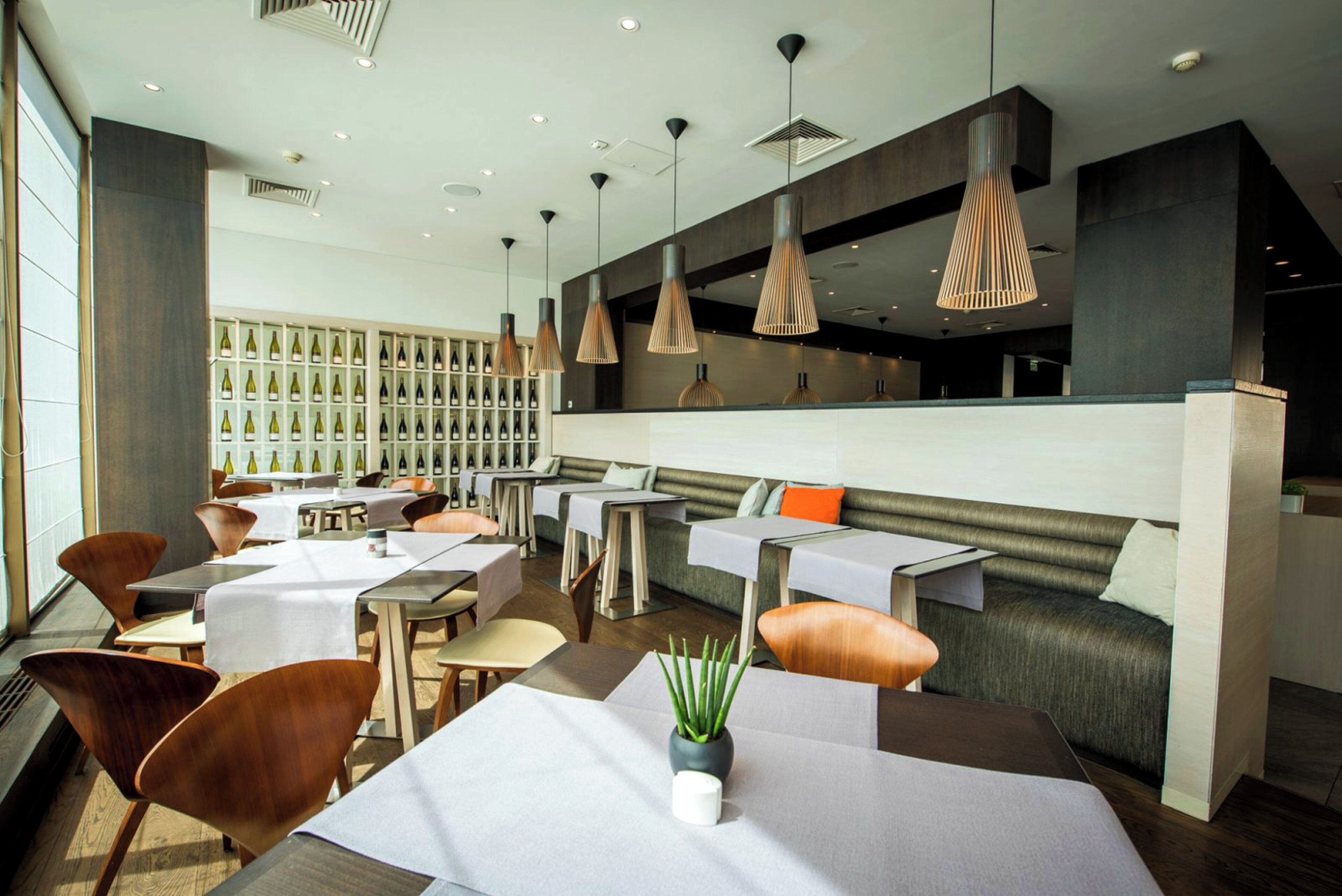 A modern restaurant interior with multiple dining tables, wooden chairs, pendant lighting, and a wine display wall.