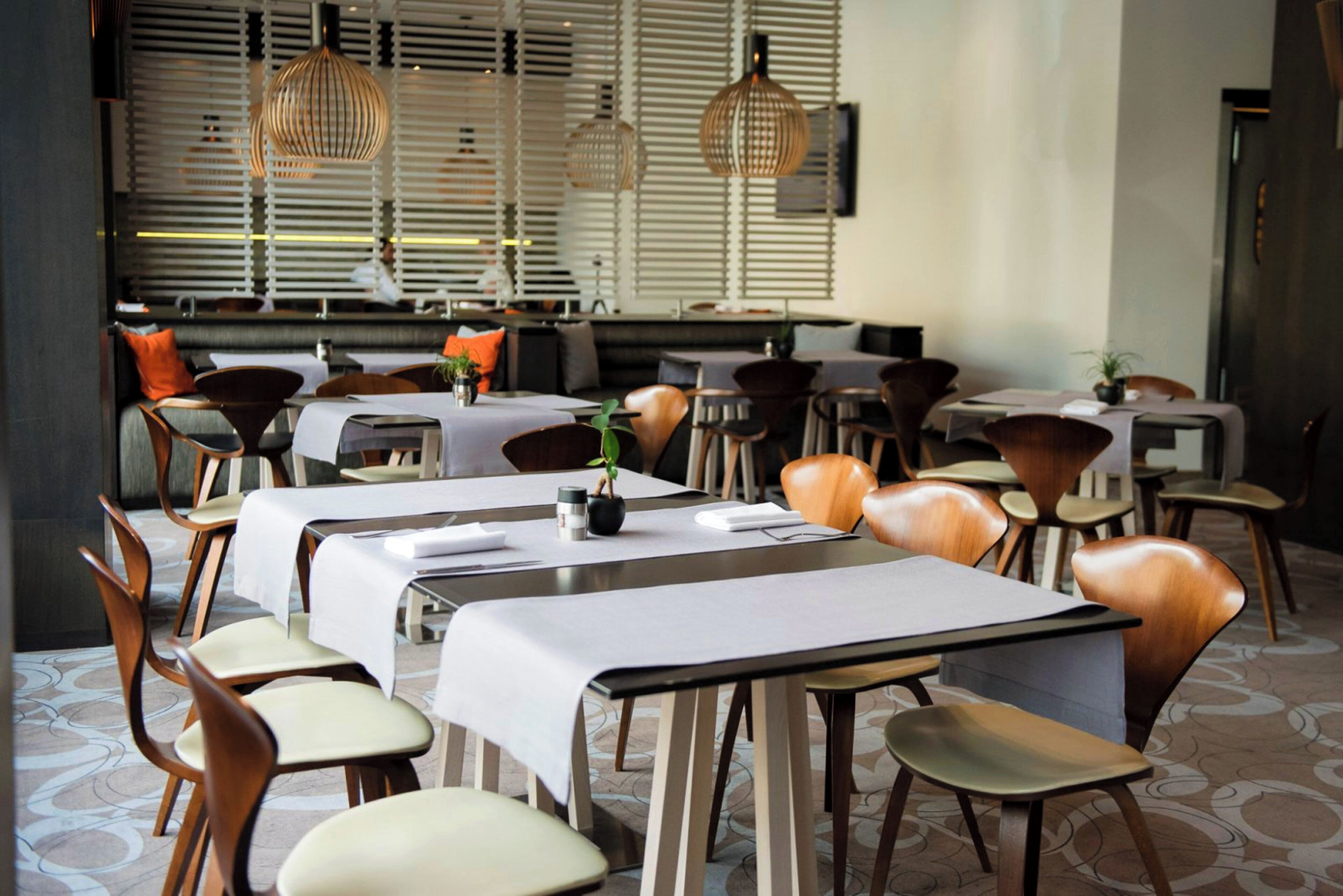 Un restaurante con varias mesas y sillas, ventanas amplias con persianas y lámparas colgantes.