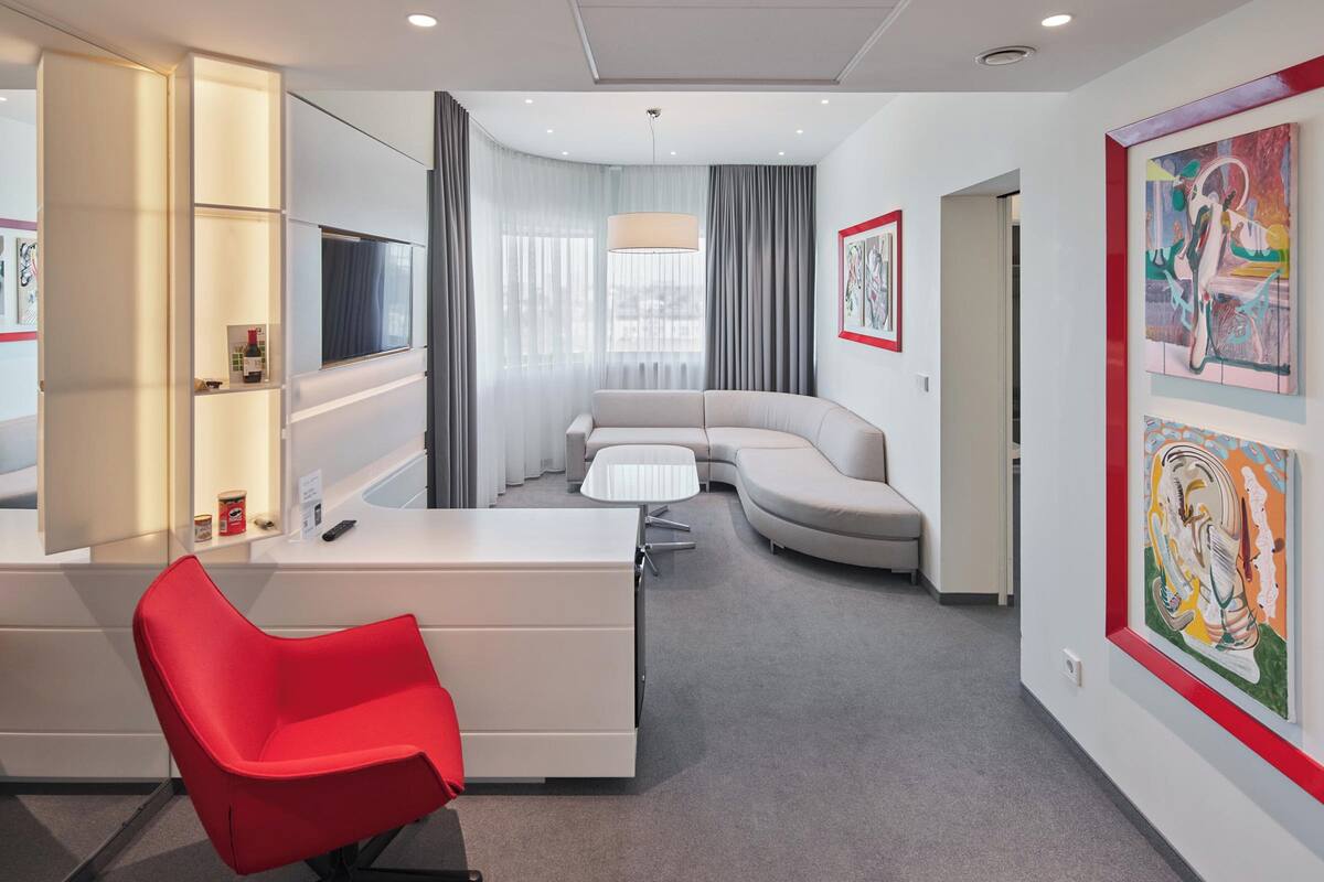 A modern hotel room with a curved sofa, a red chair, a desk with a TV, and artwork on the walls.