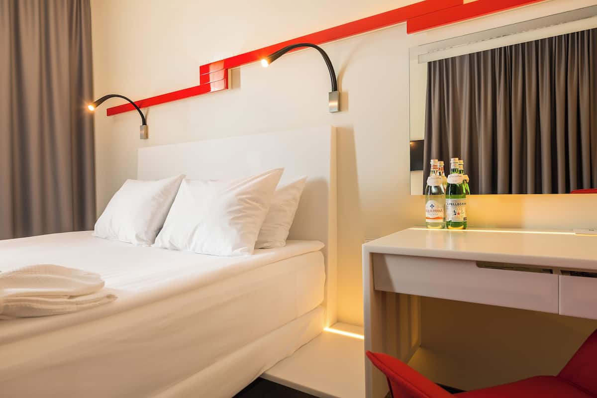 A hotel room with a bed, a desk, and a mirror. There are bottles on the desk.
