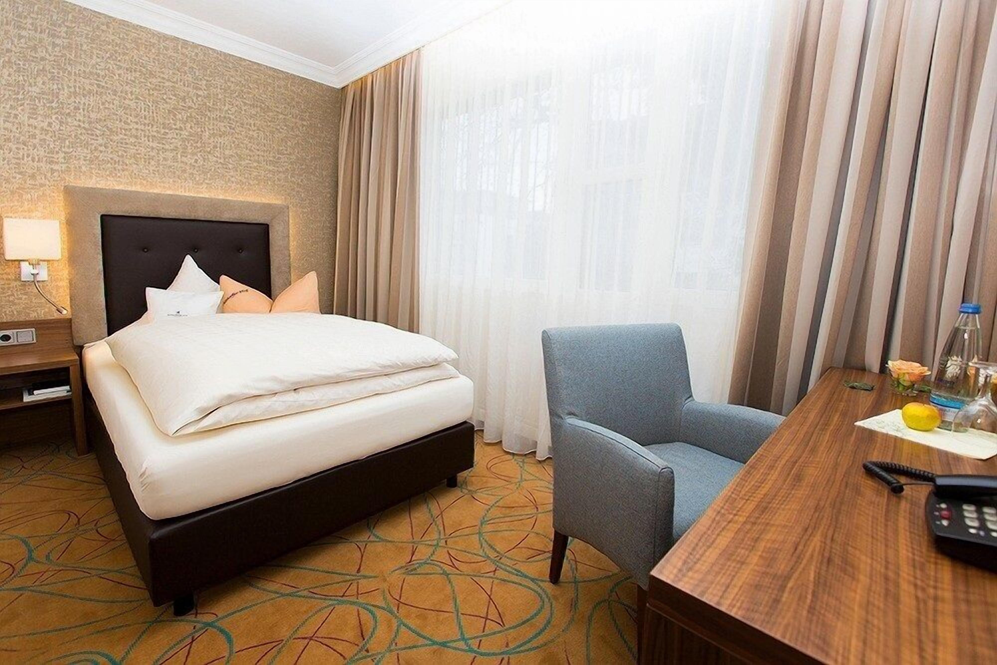 Habitación de hotel con cama, escritorio, silla, lámpara y ventana con cortinas.