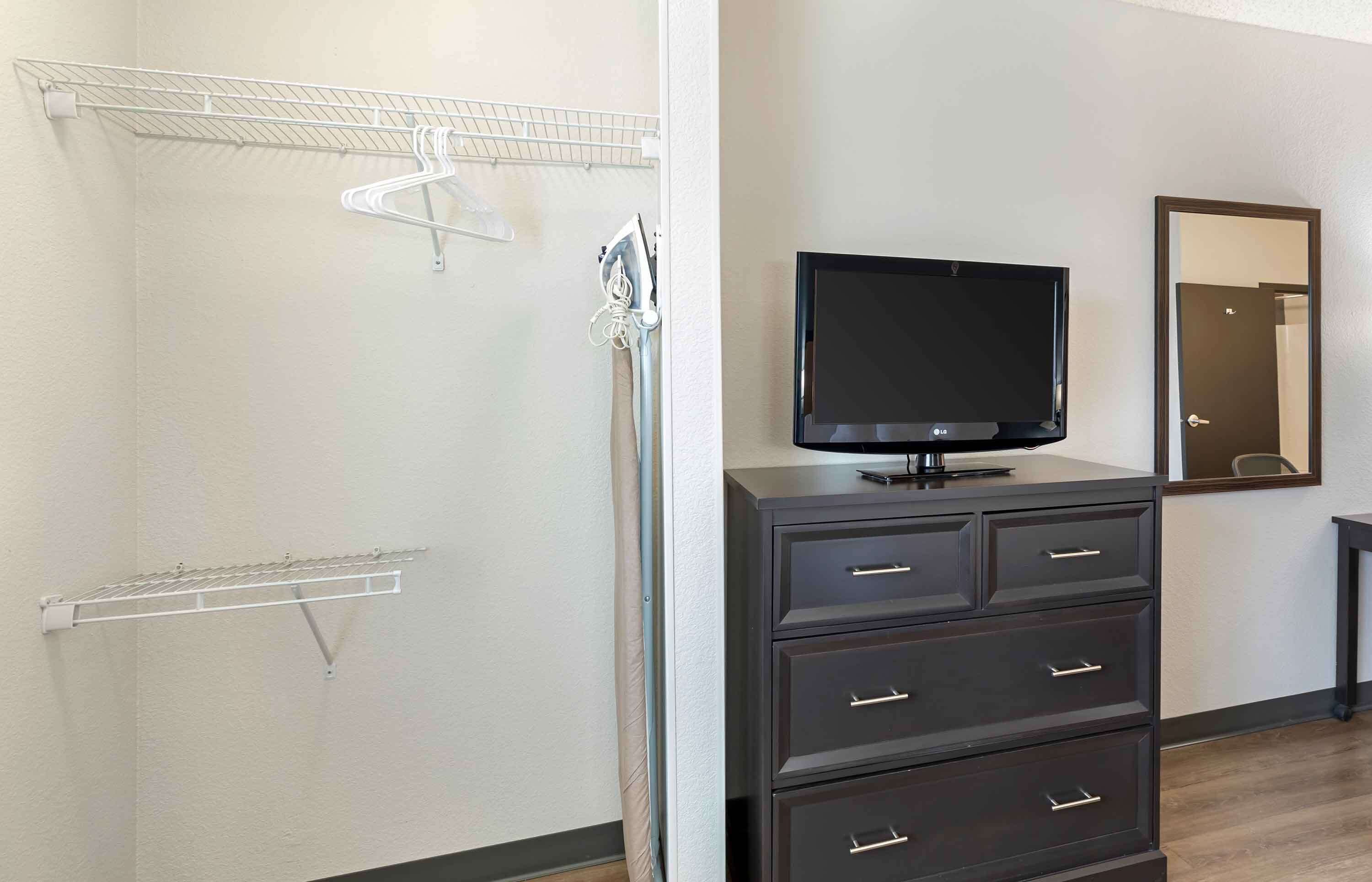 A room with a dark-colored dresser, a flat-screen TV, a mirror, and a metal drying rack on the wall.