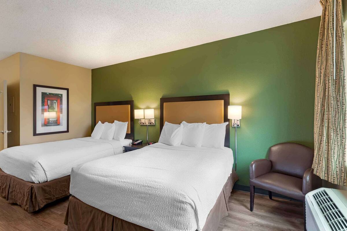 A hotel room with two beds, a chair, and a wall-mounted air conditioner.