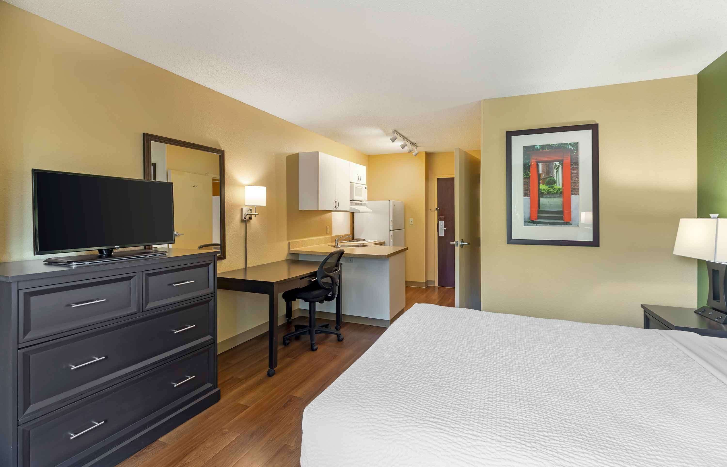 A hotel room with a bed, a desk with a chair, a television on a dresser, a microwave, and a refrigerator.