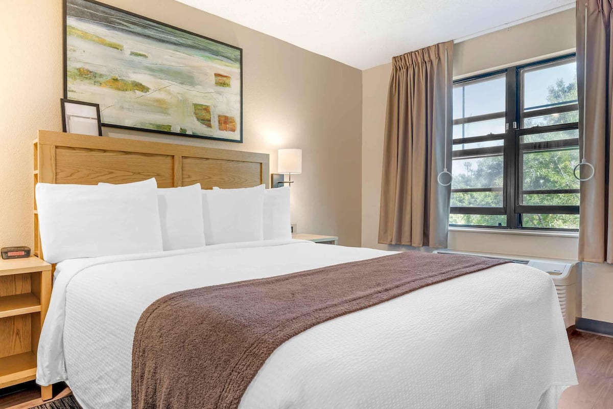 Een hotelkamer met een groot bed, hoofdeinde, nachtkastje met lamp, raam met gordijnen en een schilderij aan de muur.