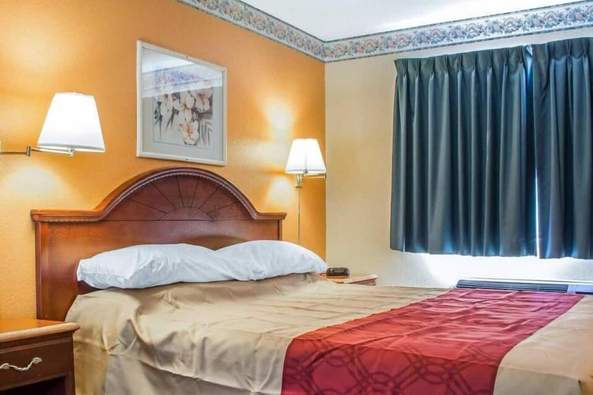 A hotel room with a bed, two bedside lamps, a nightstand, a framed picture on the wall, and a window with curtains.