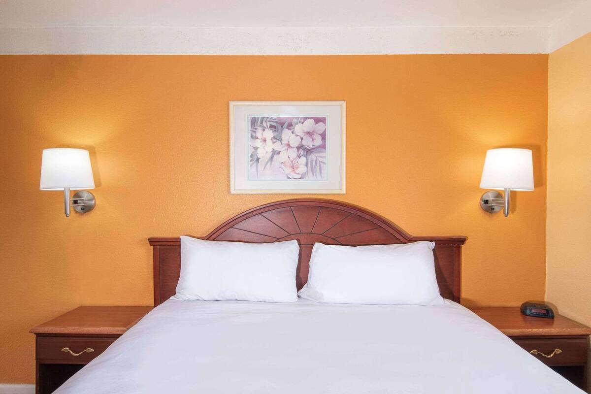 A hotel room with a bed, two bedside lamps, a nightstand with a clock, and a framed picture on the wall.