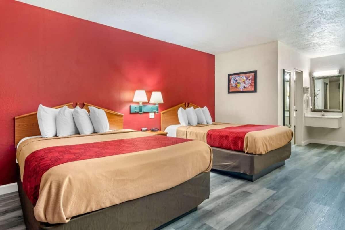 A hotel room with two beds, a red accent wall, a painting, and a mirror.