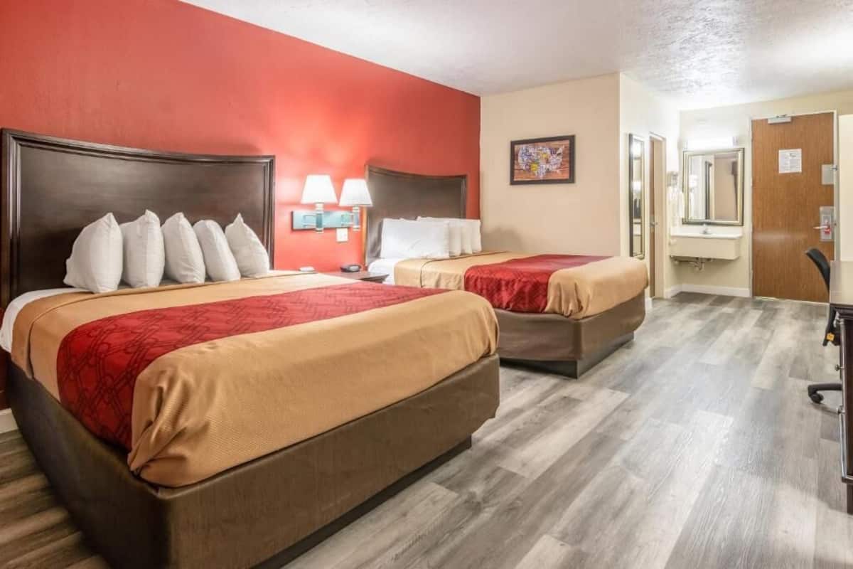 A hotel room with two beds, a wooden floor, a red accent wall, a painting, and a bathroom door.
