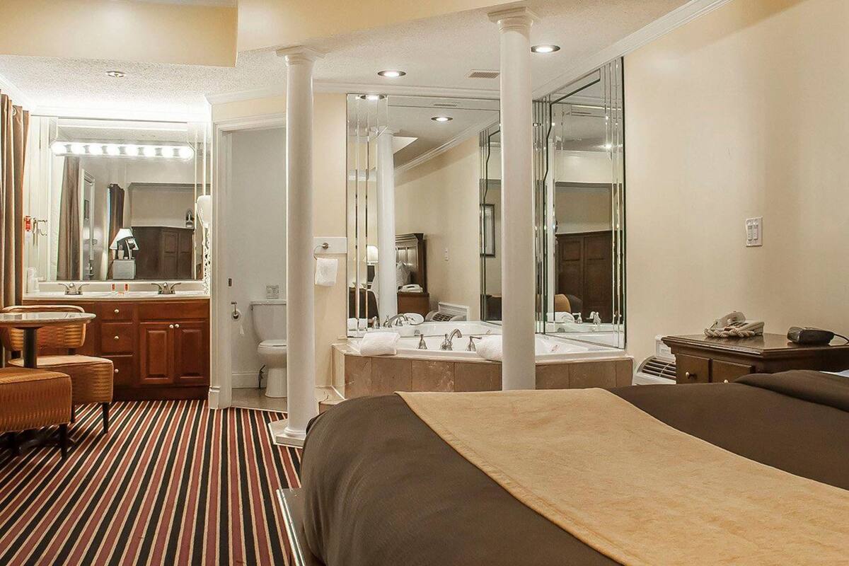 A hotel room with a large bed, a sitting area with a chair and a small table, a bathroom with a large mirror, and a striped carpet.
