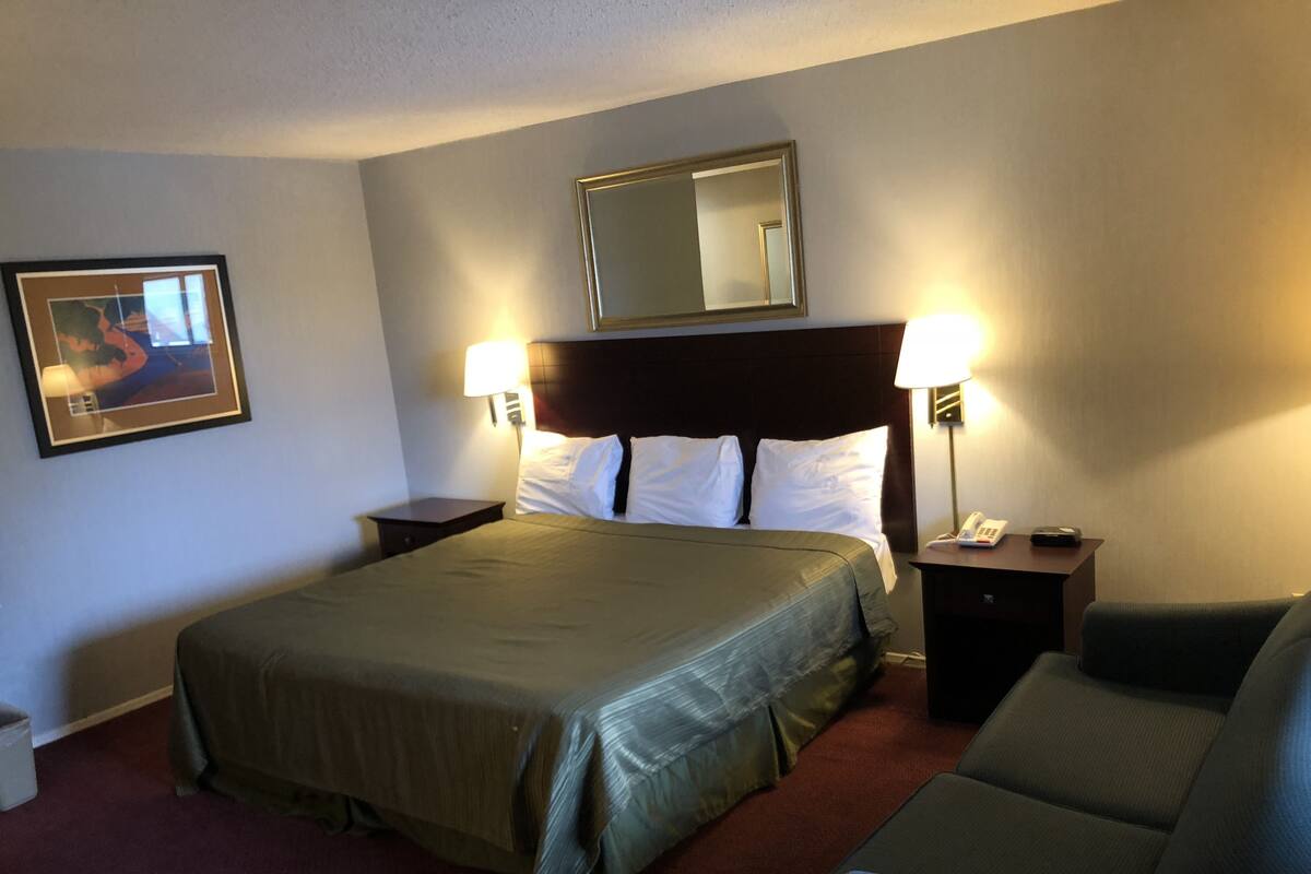 A hotel room with a large bed, two bedside tables with lamps, a chair, a framed picture on the wall, and a mirror above the bed.