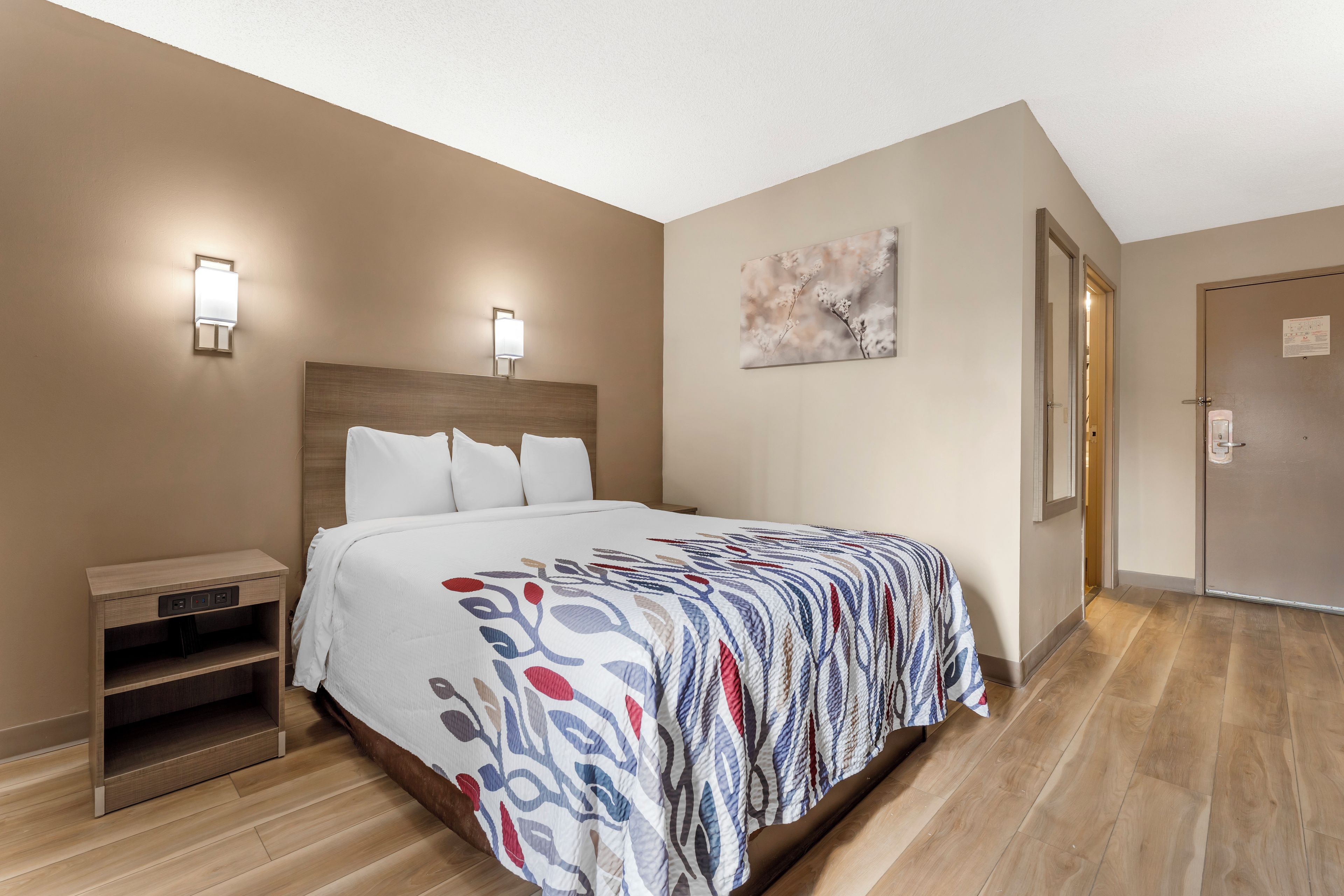 Habitación de hotel con cama, mesitas de noche, una puerta y una lámpara fijada a la pared.