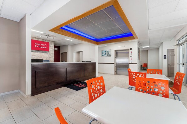 A modern restaurant with a reception area, a counter, and a dining area with orange chairs and a white table. - Red Roof Inn Johnstown (Johnstown)