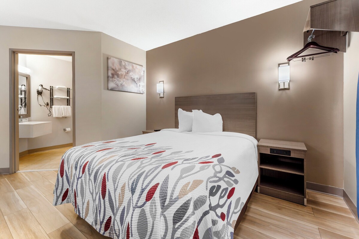 A modern hotel room with a large bed, a bedside table, a nightstand, a wall-mounted light, and a bathroom visible through an open door.