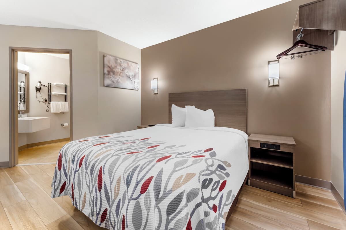 Ein modernes Hotelzimmer mit einem großen Bett, einem Nachttisch, einer Wandleuchte und einem durchgehenden Türrahmen einsehbaren Badezimmer.
