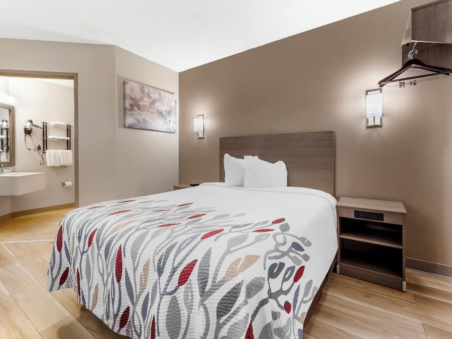 A hotel room with a large bed, a bedside table, a nightstand, a wall-mounted lamp, and a bathroom visible through an open door.