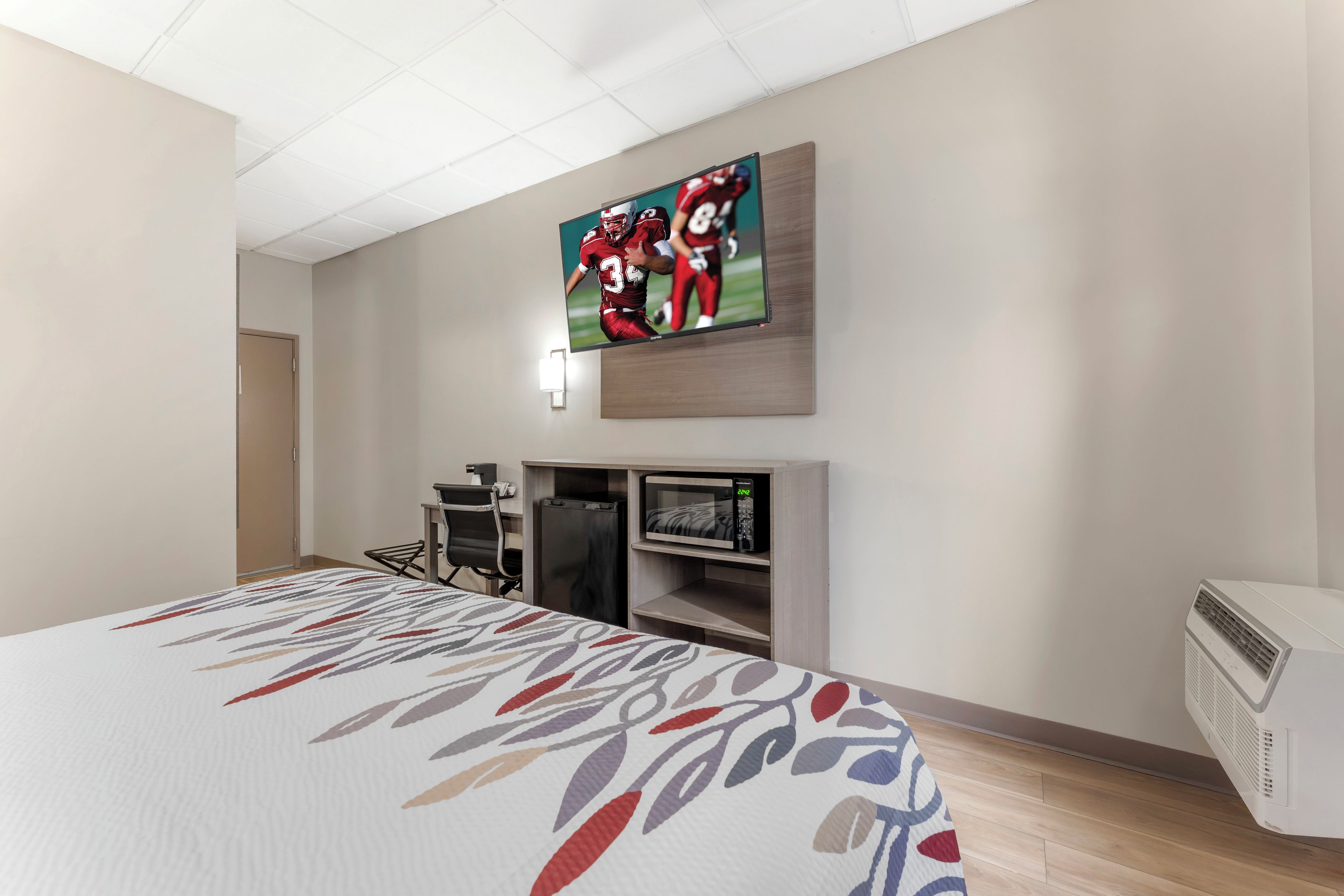 A hotel room with a bed, a flat-screen TV mounted on the wall, a desk with a chair, a microwave, and a portable air conditioner.