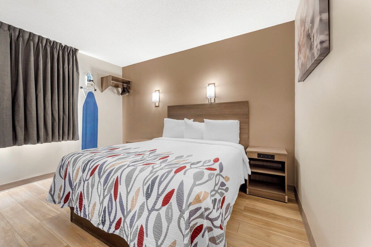 A hotel room with a bed, a nightstand, a wall-mounted shelf, and a window with curtains.