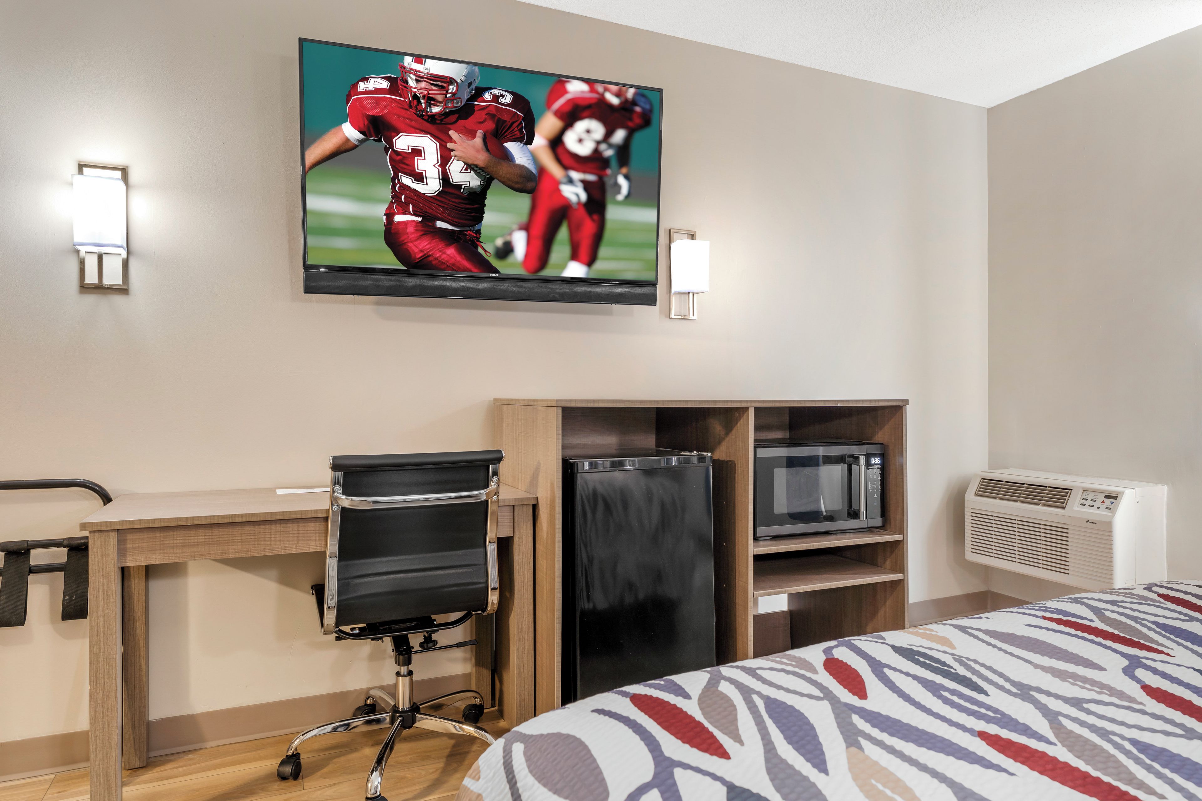 Habitación de hotel con cama, escritorio con silla, televisor montado en la pared y aire acondicionado integrado.