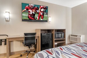 A hotel room with a bed, a desk with a chair, a television mounted on the wall, and a built-in air conditioning unit.
