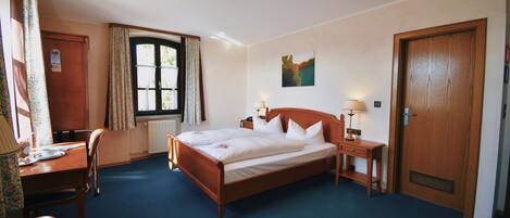 A hotel room with a wooden bed, a desk with a chair, a window with curtains, a door, and a painting on the wall.