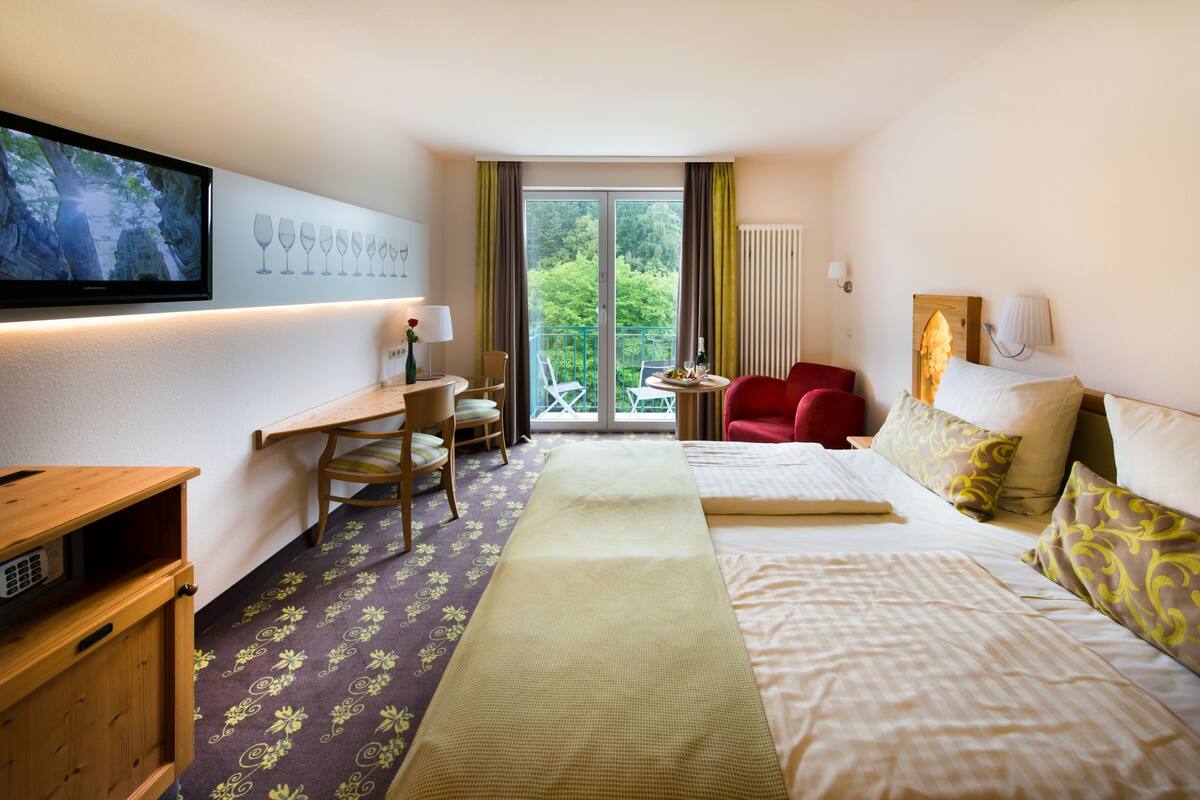 A hotel room with a bed, a desk, a chair, a TV, and a balcony with a view of trees.