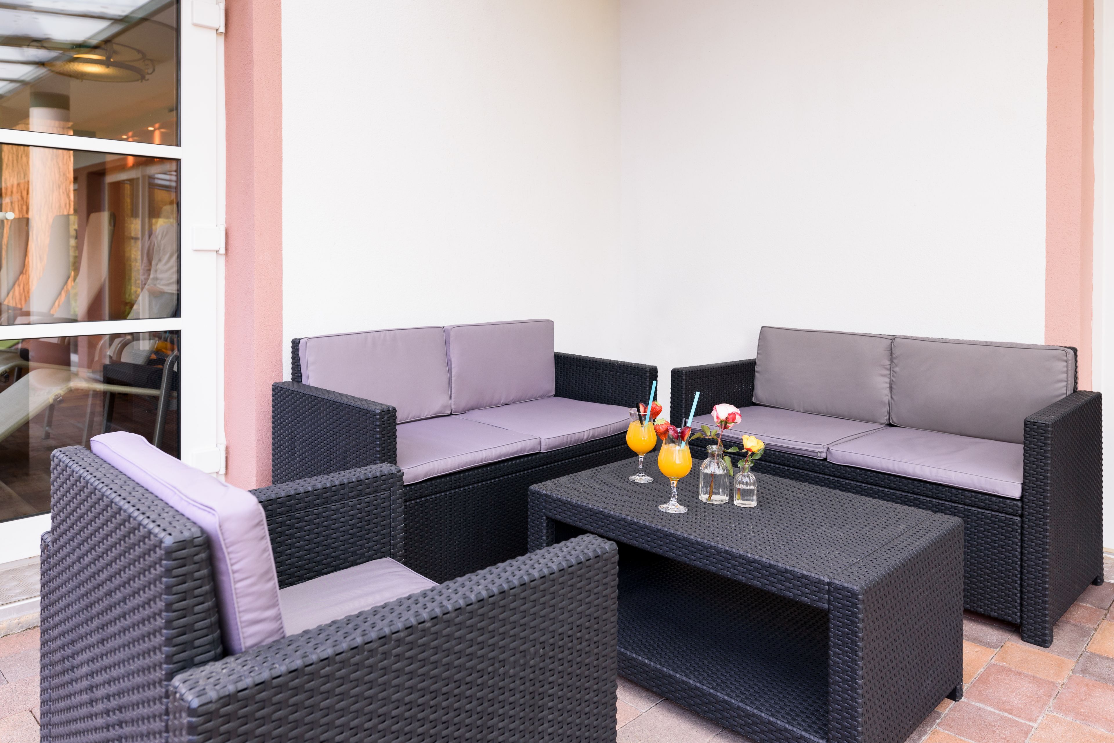 A patio area with wicker furniture, including a sofa and two chairs, and a coffee table with drinks and flowers.