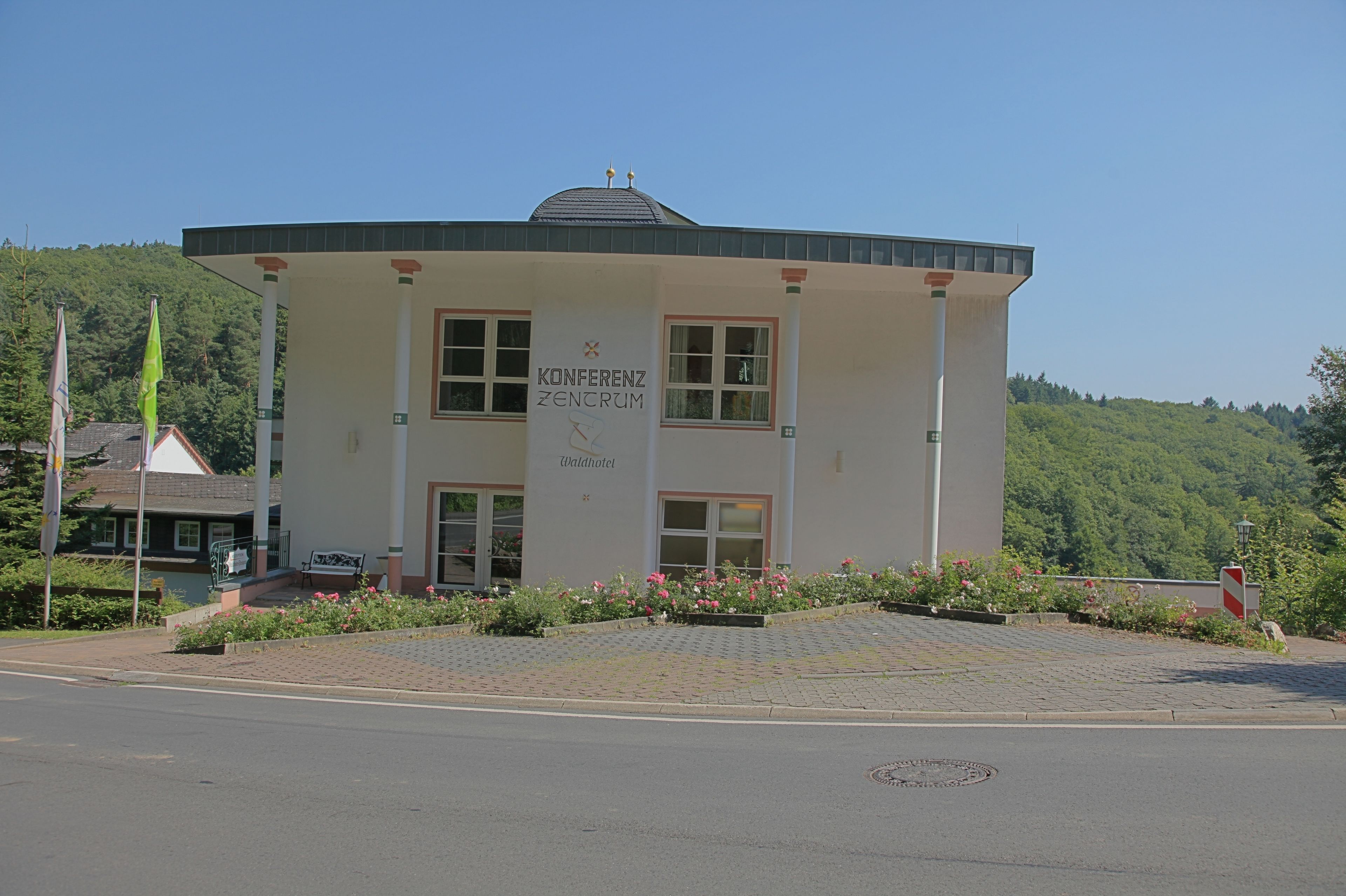 A building labeled "Konferenzzentrum Zentrums" with a flat roof, large windows, and a paved entrance area with flower beds.