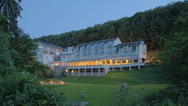 Um hotel grande com vários andares, um gramado bem cuidado e uma encosta arborizada ao fundo.